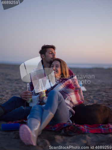 Image of Loving Young Couple Sitting On The Beach beside Campfire drinkin