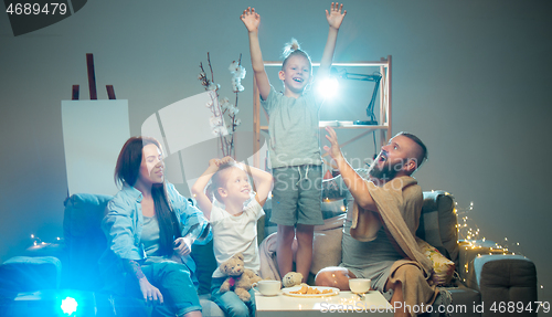 Image of Happy family watching projector, TV, movies with popcorn in the evening at home. Mother, father and kids spending time together.