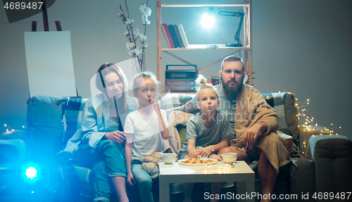 Image of Happy family watching projector, TV, movies with popcorn in the evening at home. Mother, father and kids spending time together.