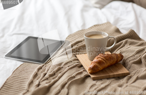 Image of tablet pc, coffee cup and croissant on bed at home