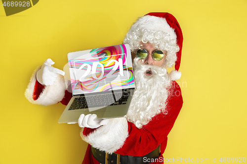 Image of Stylish Santa Claus in traditional costume with modern device pointing on 2021 on yellow studio background