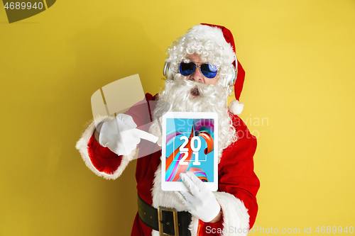 Image of Stylish Santa Claus in traditional costume with modern device pointing on 2021 on yellow studio background