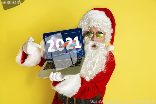 Image of Stylish Santa Claus in traditional costume with modern device pointing on 2021 on yellow studio background