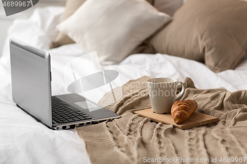 Image of laptop, cup of coffee and croissant on bed at home