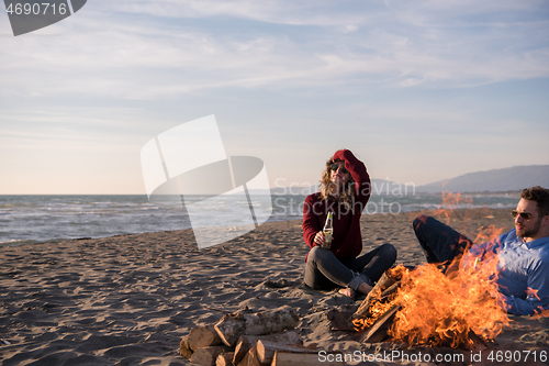 Image of Young Couple Sitting On The Beach beside Campfire drinking beer