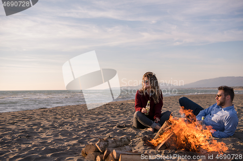 Image of Young Couple Sitting On The Beach beside Campfire drinking beer