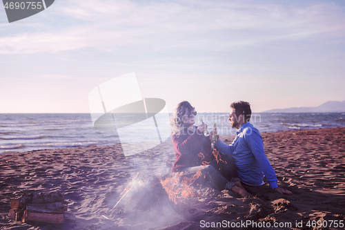 Image of Young Couple Sitting On The Beach beside Campfire drinking beer