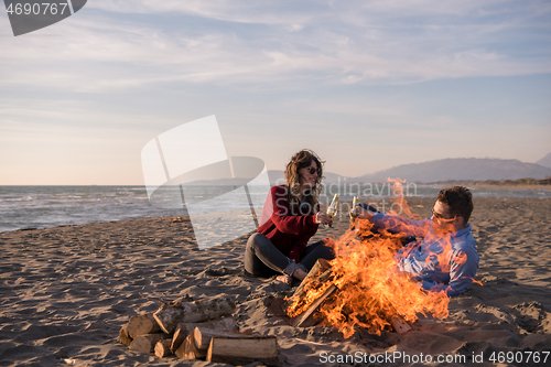 Image of Young Couple Sitting On The Beach beside Campfire drinking beer