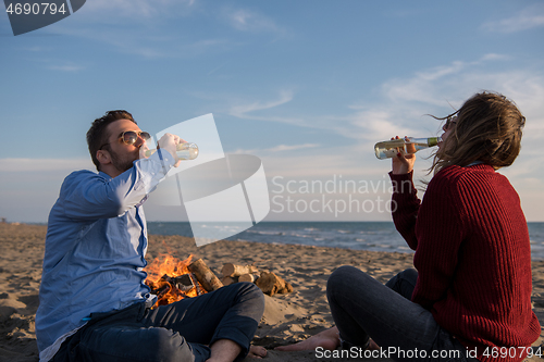 Image of Young Couple Sitting On The Beach beside Campfire drinking beer