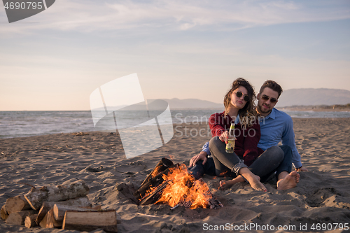 Image of Young Couple Sitting On The Beach beside Campfire drinking beer