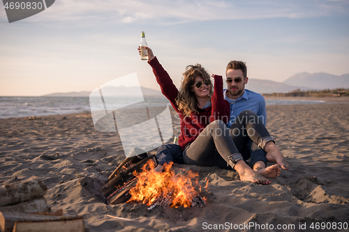 Image of Young Couple Sitting On The Beach beside Campfire drinking beer