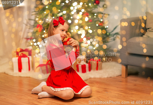 Image of girl in red dress hugging teddy bear at home