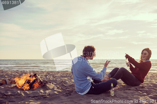 Image of Young Couple Sitting On The Beach beside Campfire drinking beer