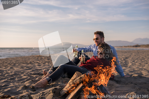 Image of Young Couple Sitting On The Beach beside Campfire drinking beer