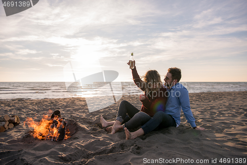 Image of Young Couple Sitting On The Beach beside Campfire drinking beer