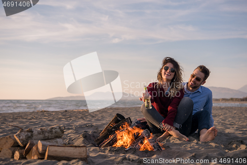 Image of Young Couple Sitting On The Beach beside Campfire drinking beer