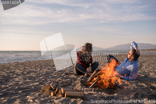 Image of Young Couple Sitting On The Beach beside Campfire drinking beer