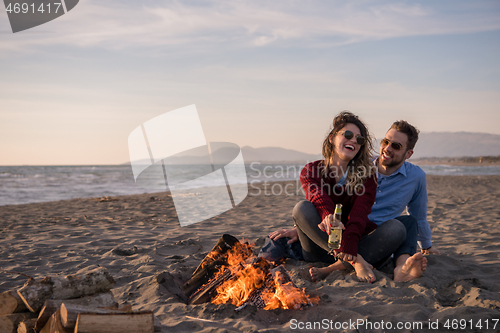 Image of Young Couple Sitting On The Beach beside Campfire drinking beer
