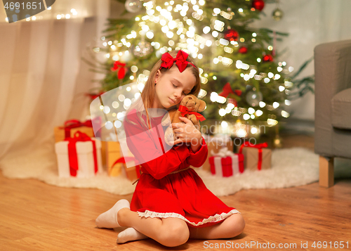 Image of girl in red dress hugging teddy bear at home