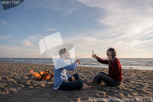 Image of Young Couple Sitting On The Beach beside Campfire drinking beer