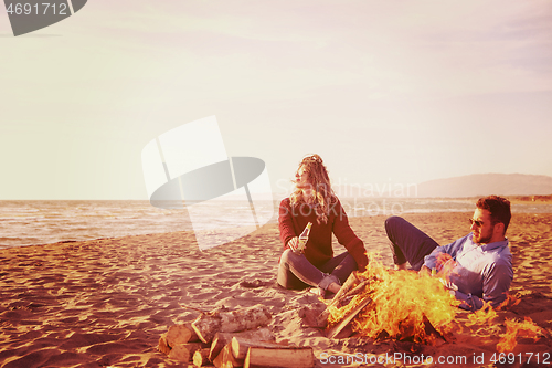 Image of Young Couple Sitting On The Beach beside Campfire drinking beer