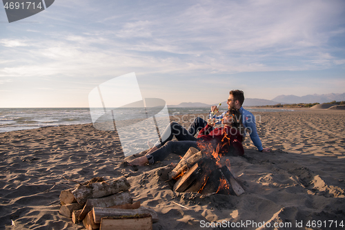 Image of Young Couple Sitting On The Beach beside Campfire drinking beer