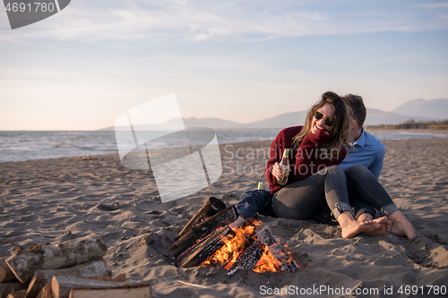 Image of Young Couple Sitting On The Beach beside Campfire drinking beer