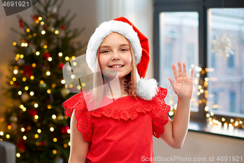 Image of happy girl in santa helper hat waving hand at home