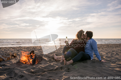 Image of Young Couple Sitting On The Beach beside Campfire drinking beer