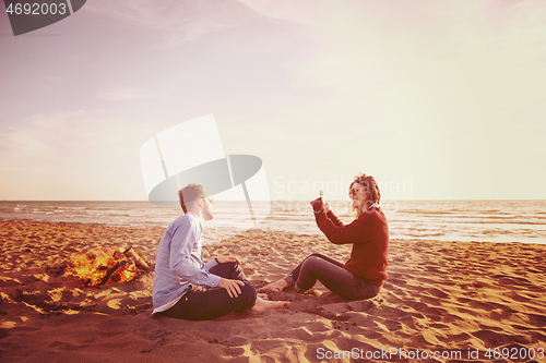 Image of Young Couple Sitting On The Beach beside Campfire drinking beer