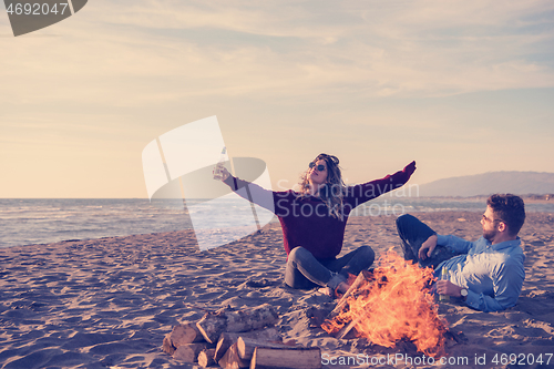 Image of Young Couple Sitting On The Beach beside Campfire drinking beer