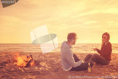 Image of Young Couple Sitting On The Beach beside Campfire drinking beer