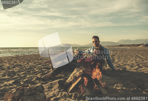 Image of Young Couple Sitting On The Beach beside Campfire drinking beer