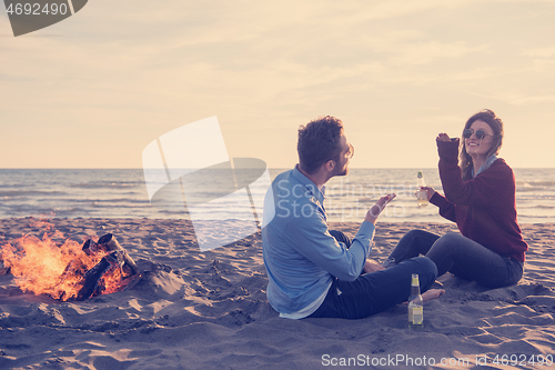 Image of Young Couple Sitting On The Beach beside Campfire drinking beer