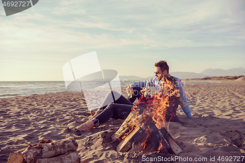 Image of Young Couple Sitting On The Beach beside Campfire drinking beer