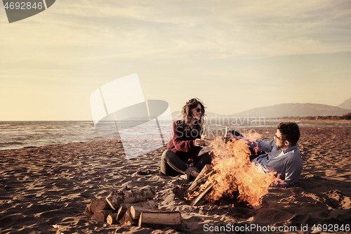 Image of Young Couple Sitting On The Beach beside Campfire drinking beer