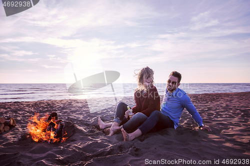 Image of Young Couple Sitting On The Beach beside Campfire drinking beer