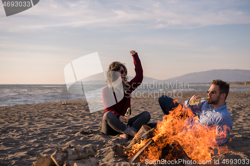 Image of Young Couple Sitting On The Beach beside Campfire drinking beer