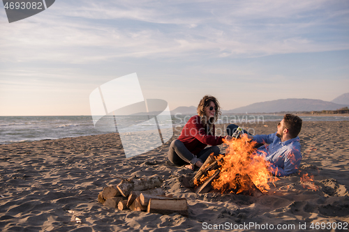 Image of Young Couple Sitting On The Beach beside Campfire drinking beer