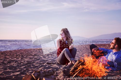 Image of Young Couple Sitting On The Beach beside Campfire drinking beer