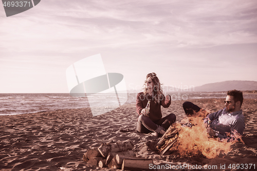 Image of Young Couple Sitting On The Beach beside Campfire drinking beer