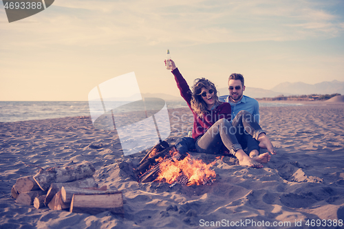 Image of Young Couple Sitting On The Beach beside Campfire drinking beer