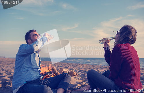 Image of Young Couple Sitting On The Beach beside Campfire drinking beer