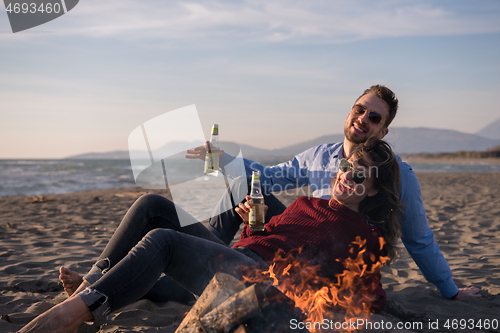 Image of Young Couple Sitting On The Beach beside Campfire drinking beer