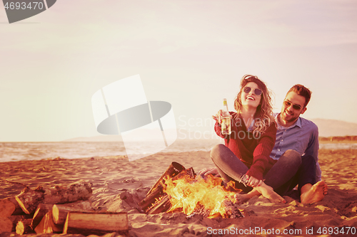 Image of Young Couple Sitting On The Beach beside Campfire drinking beer