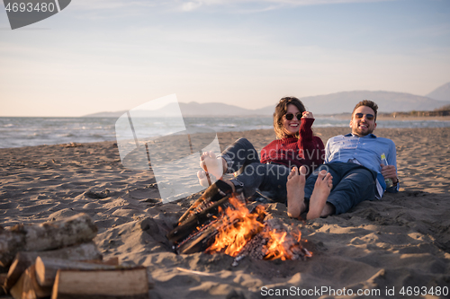 Image of Young Couple Sitting On The Beach beside Campfire drinking beer