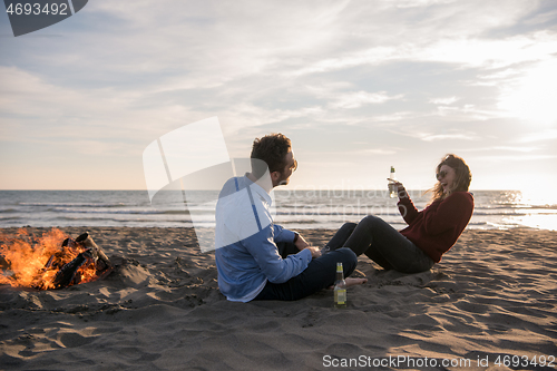 Image of Young Couple Sitting On The Beach beside Campfire drinking beer