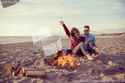 Image of Young Couple Sitting On The Beach beside Campfire drinking beer