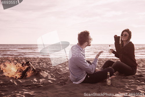 Image of Young Couple Sitting On The Beach beside Campfire drinking beer