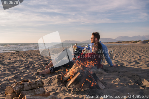 Image of Young Couple Sitting On The Beach beside Campfire drinking beer